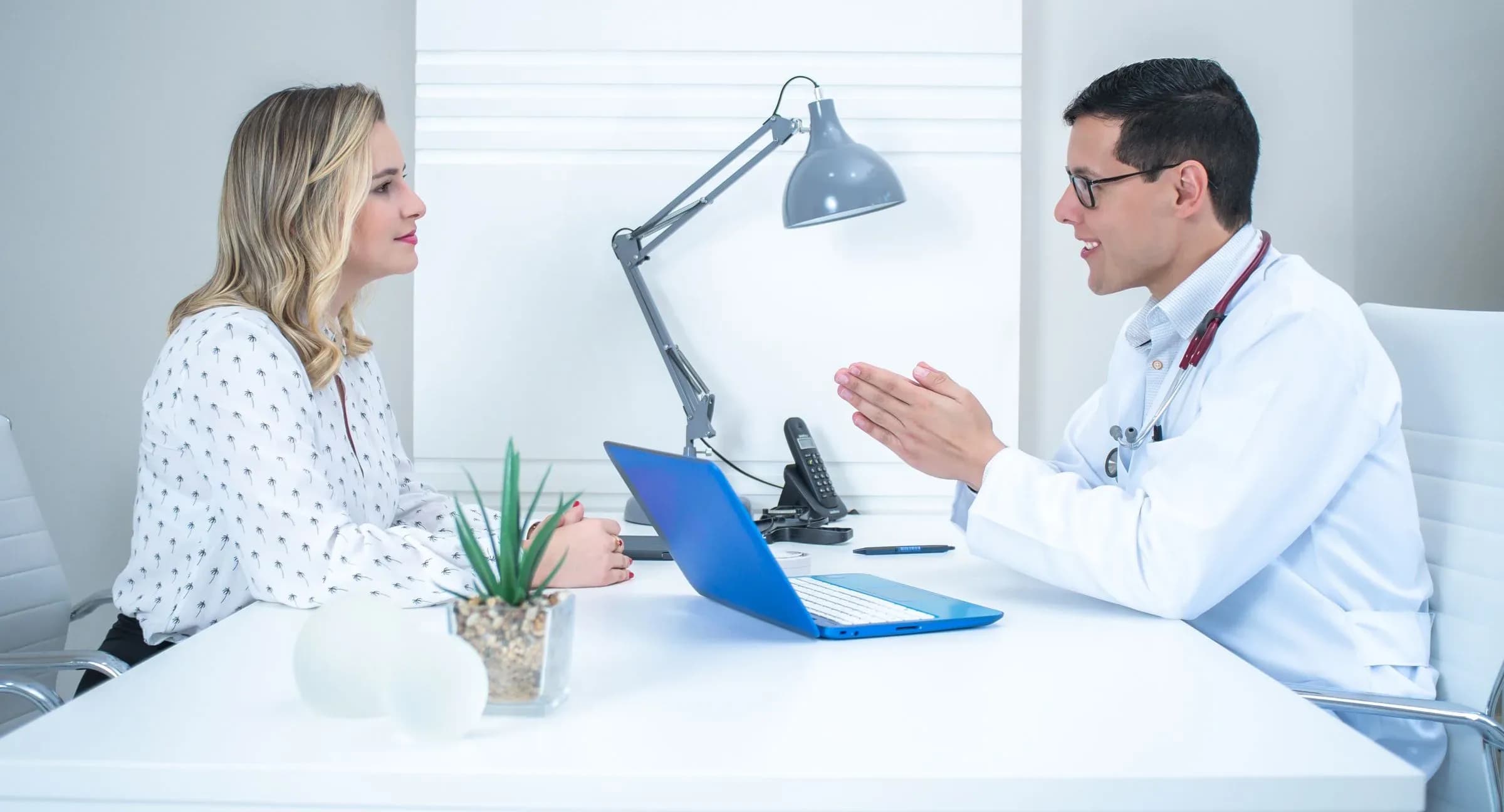 A woman and a doctor in a white coat talking across a desk in a bright consulting room.
