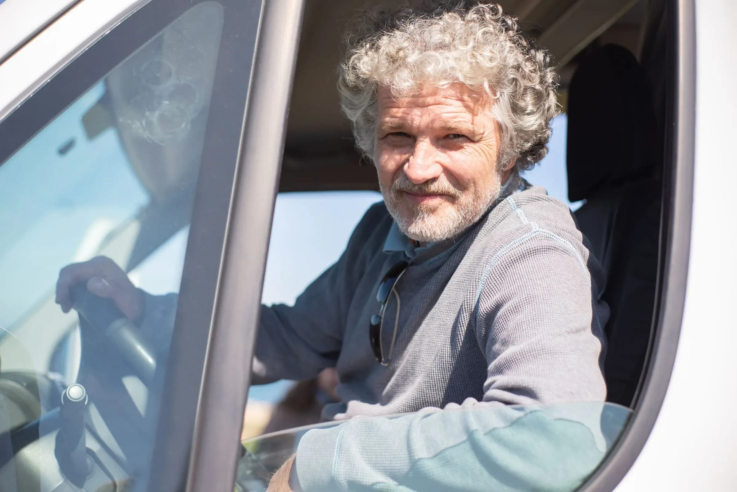 A man with grey curly hair at the wheel of a van, looking out through the open window.