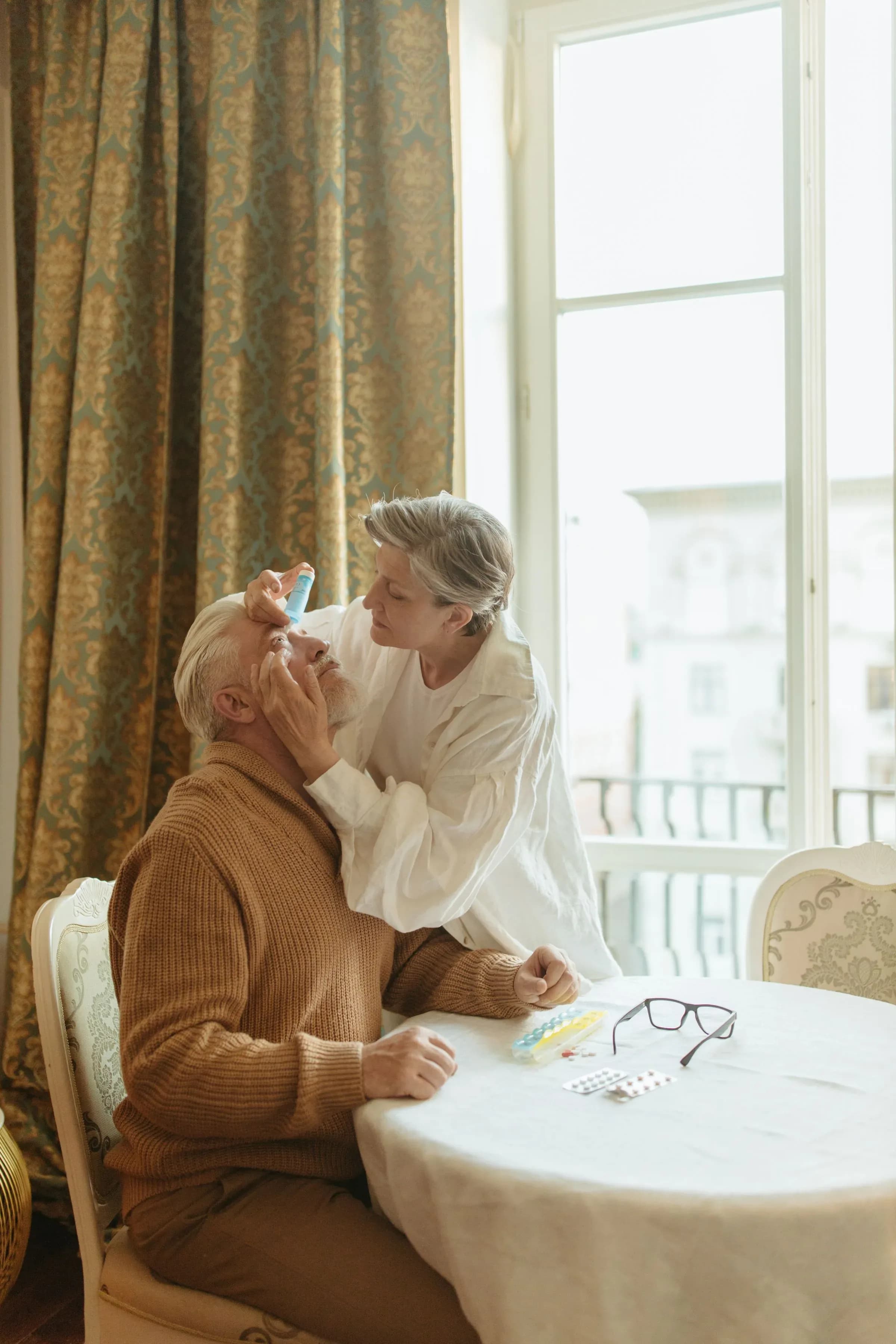 A woman putting eye drops into an older man’s eye at a table beside a window.