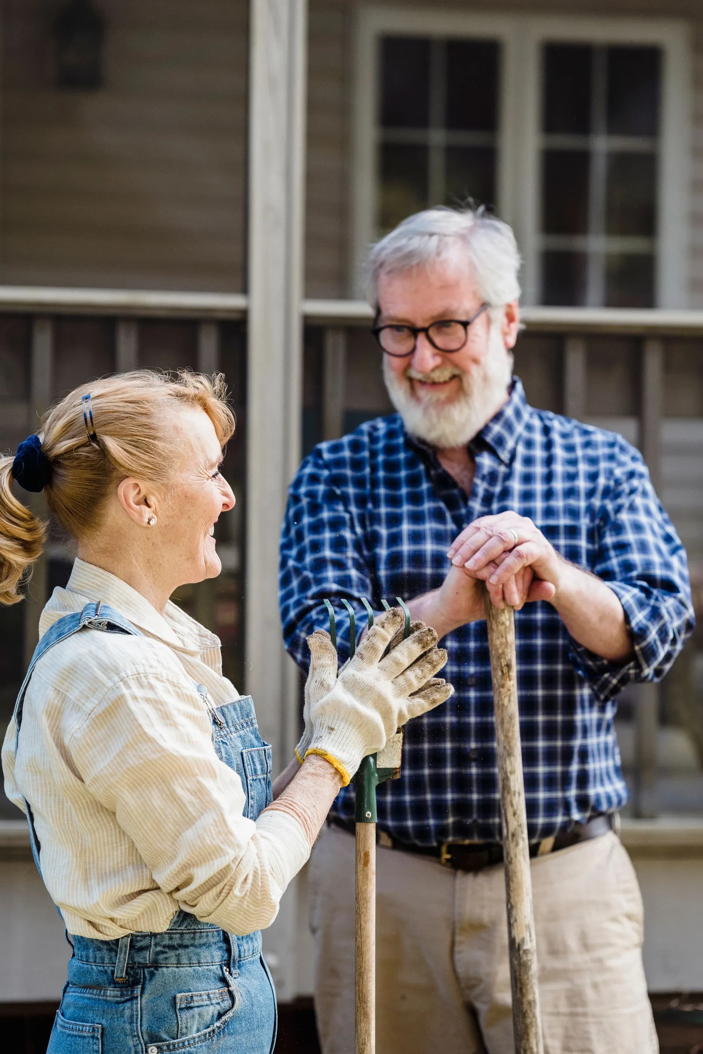 A couple in their seventies laughing together in a garden, the man in glasses leaning on a spade.
