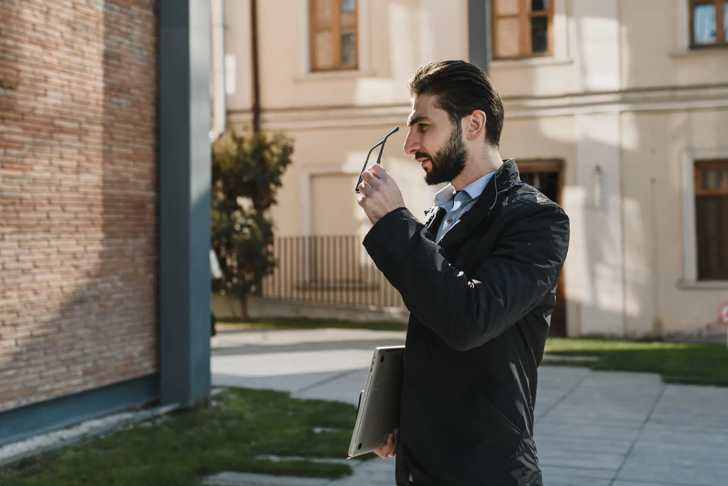 A man outdoors holding his glasses away from his face, a folder tucked under his arm.