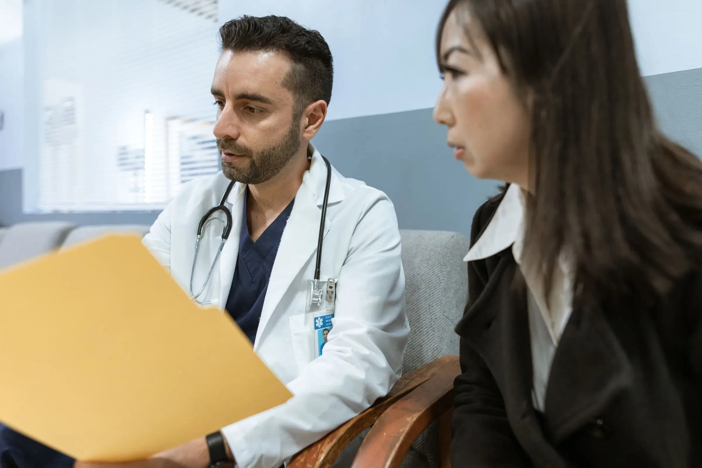 A doctor in a white coat going through notes in a folder, a patient sitting beside him.