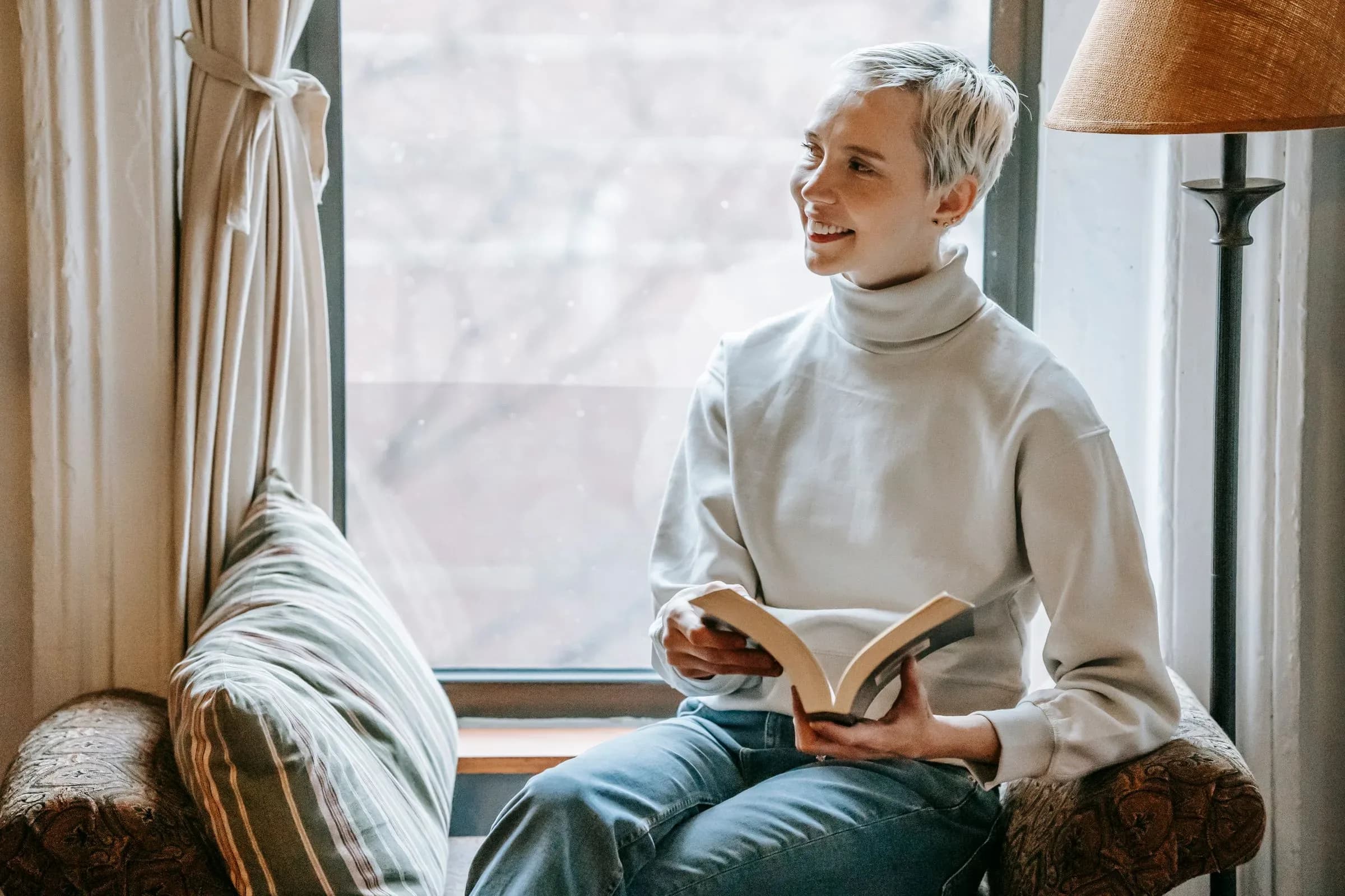 A woman with short grey hair sitting on a window seat in daylight, reading a book.