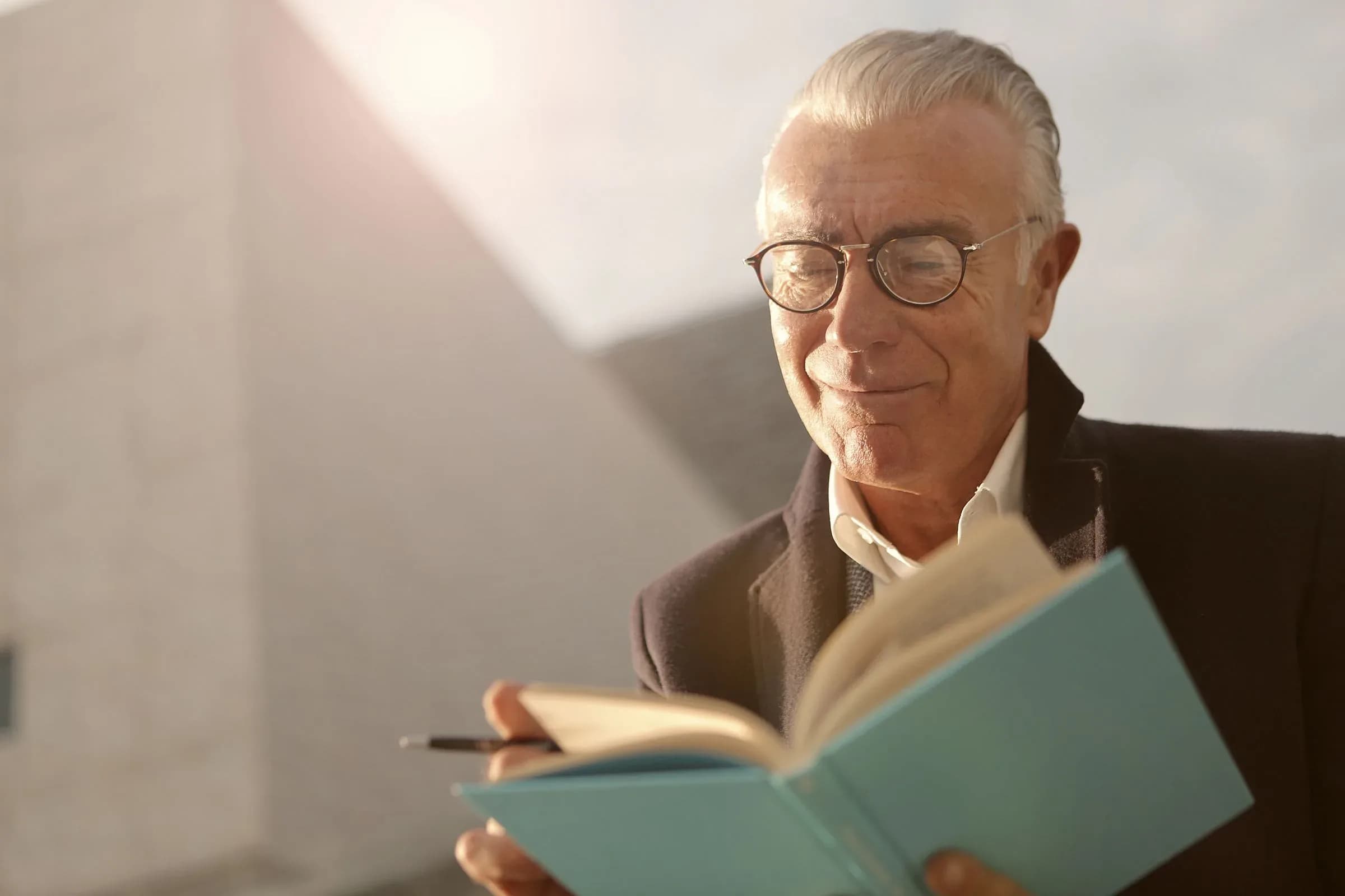 A man in his seventies in reading glasses, smiling over an open book in warm daylight.