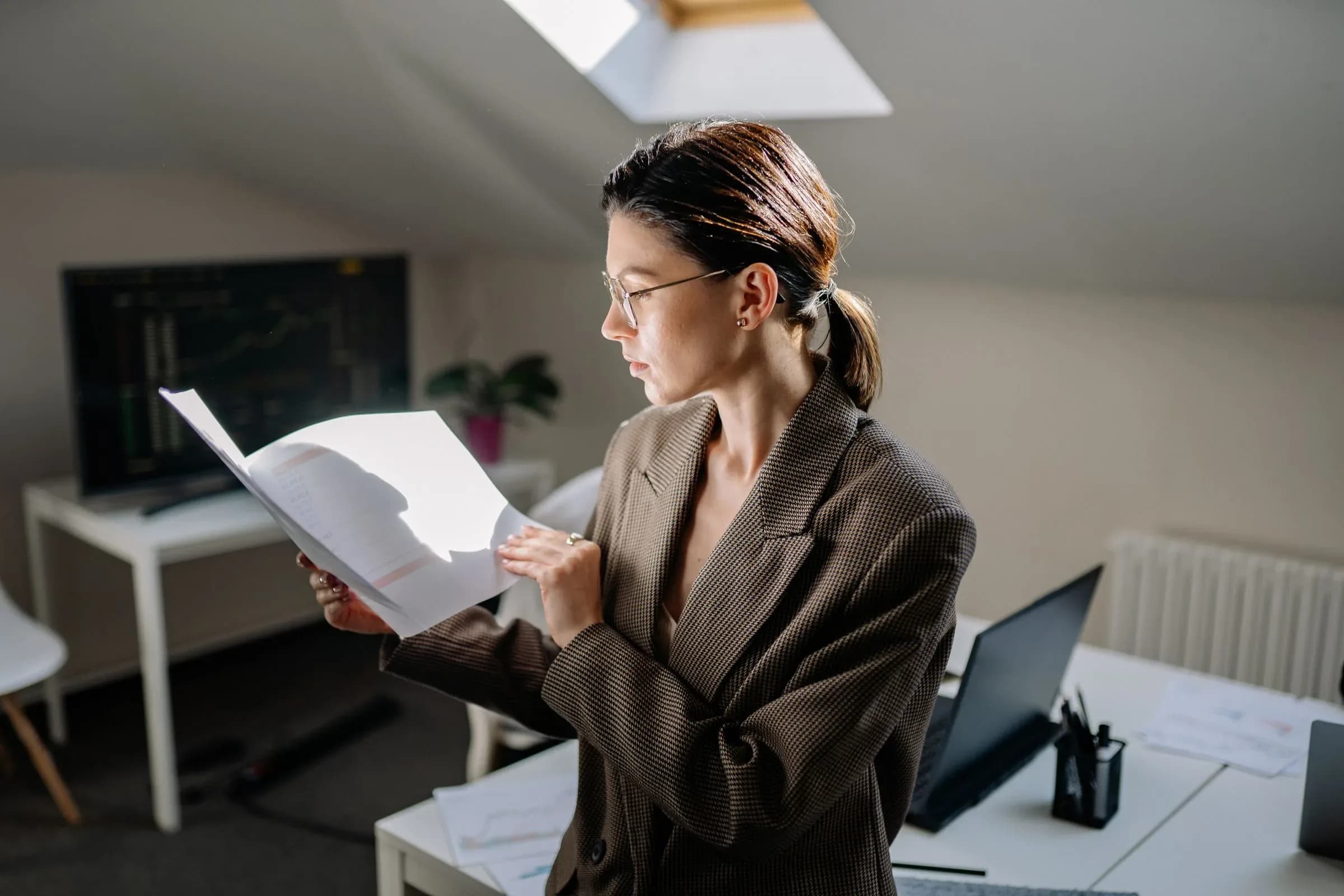 A woman in a blazer standing at a desk, reading through a printed document.