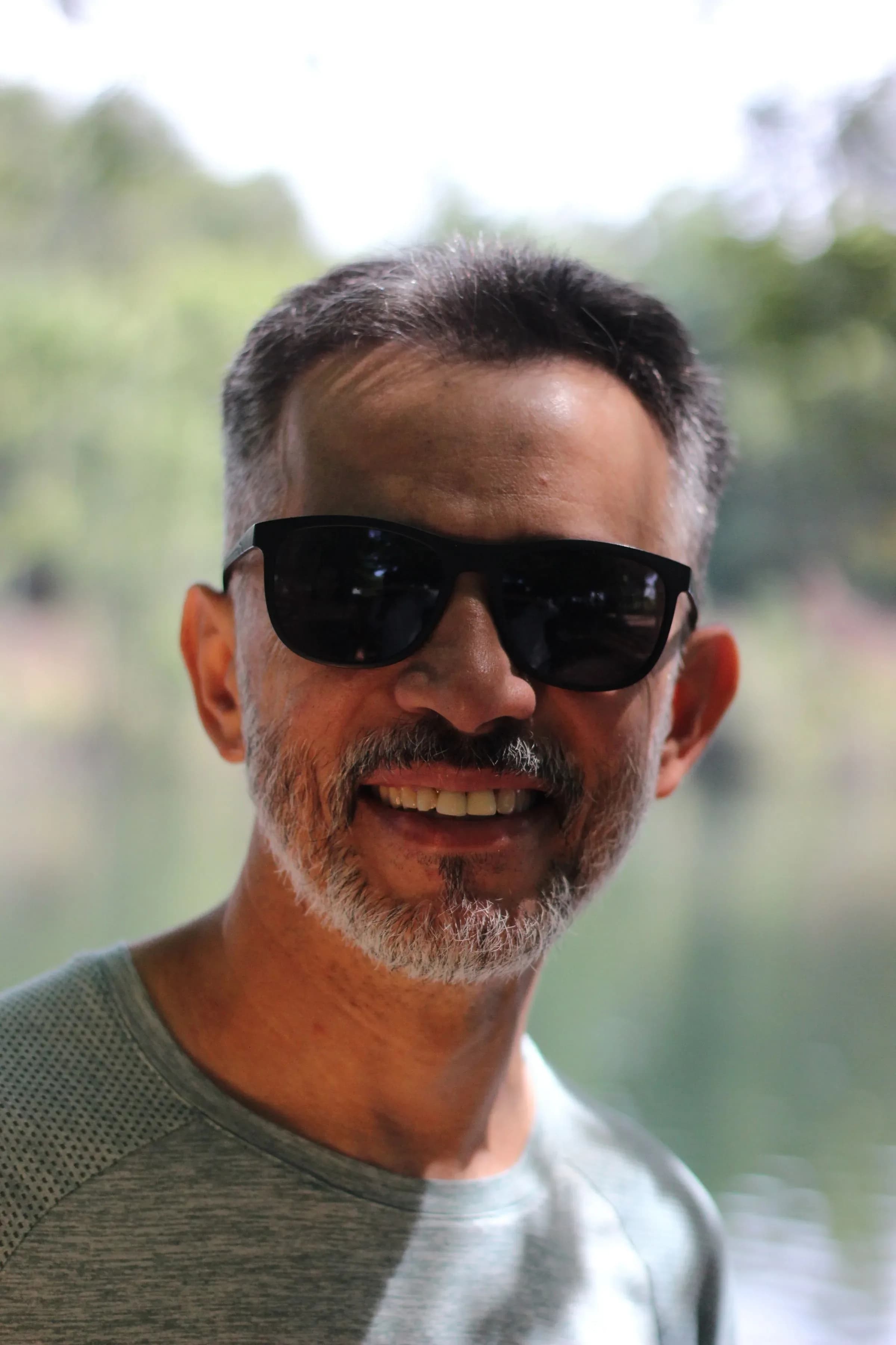 A man in his fifties with salt-and-pepper hair and sunglasses, smiling beside a lake.