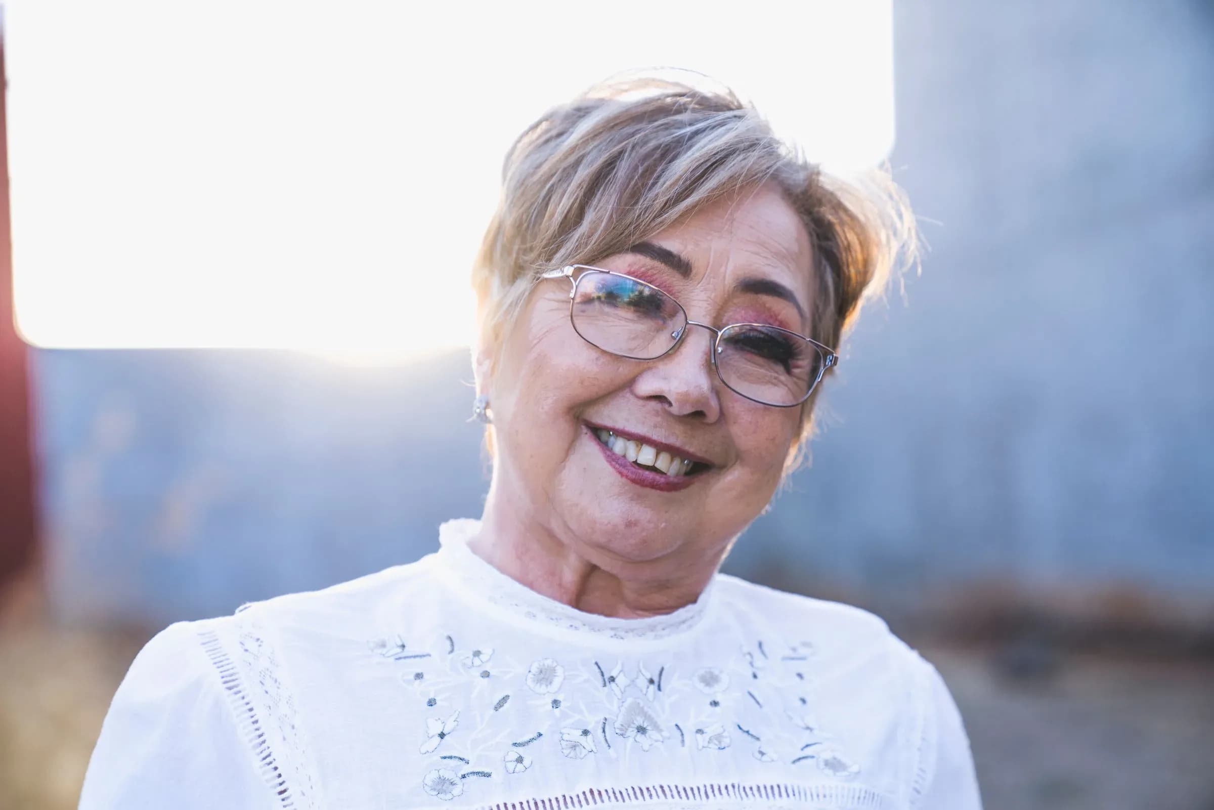 A woman in her sixties with short fair hair and wire-framed glasses, smiling in low evening sun.