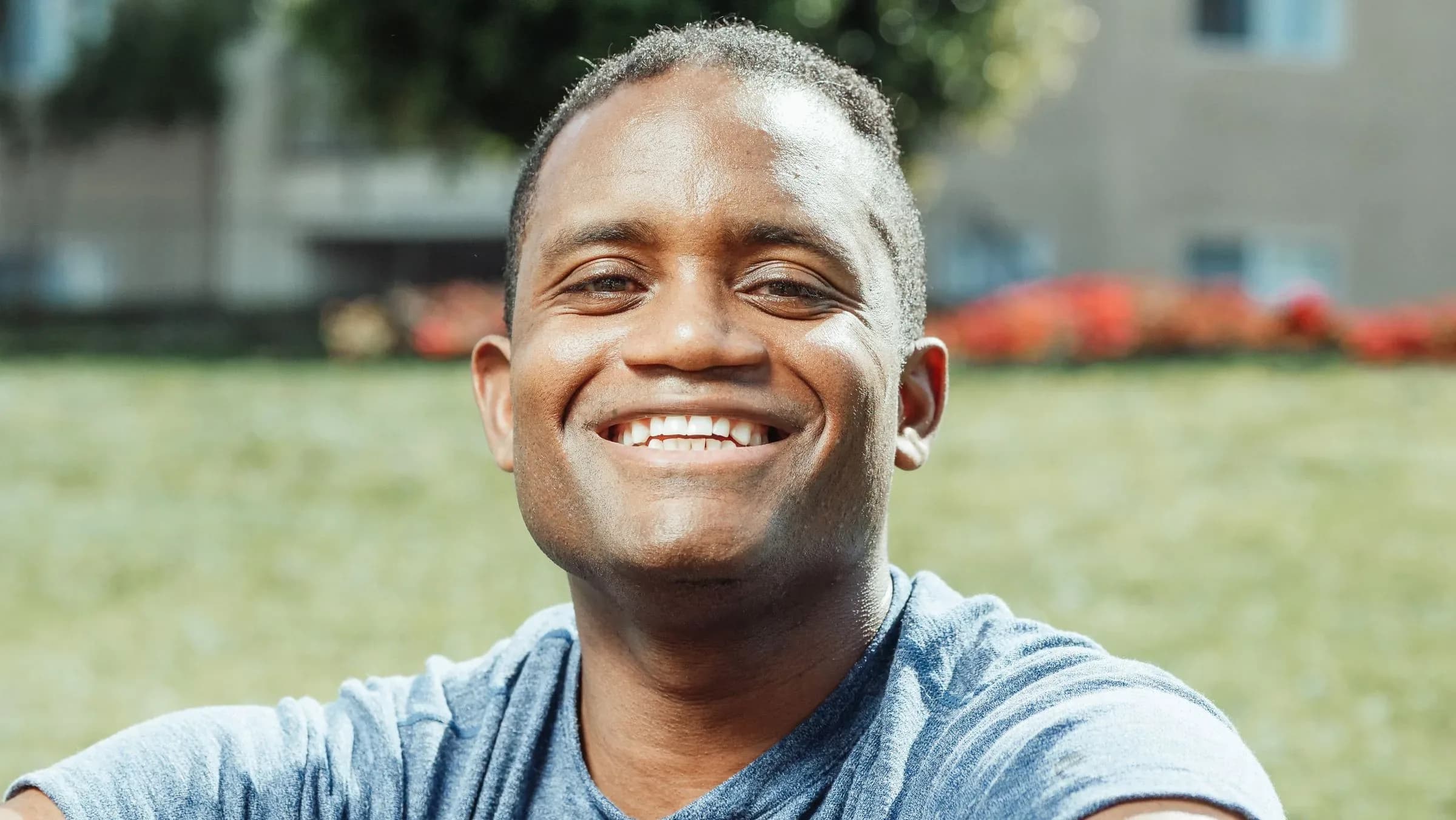 A man in his thirties sitting on the grass in a park on a sunny day, laughing at the camera.