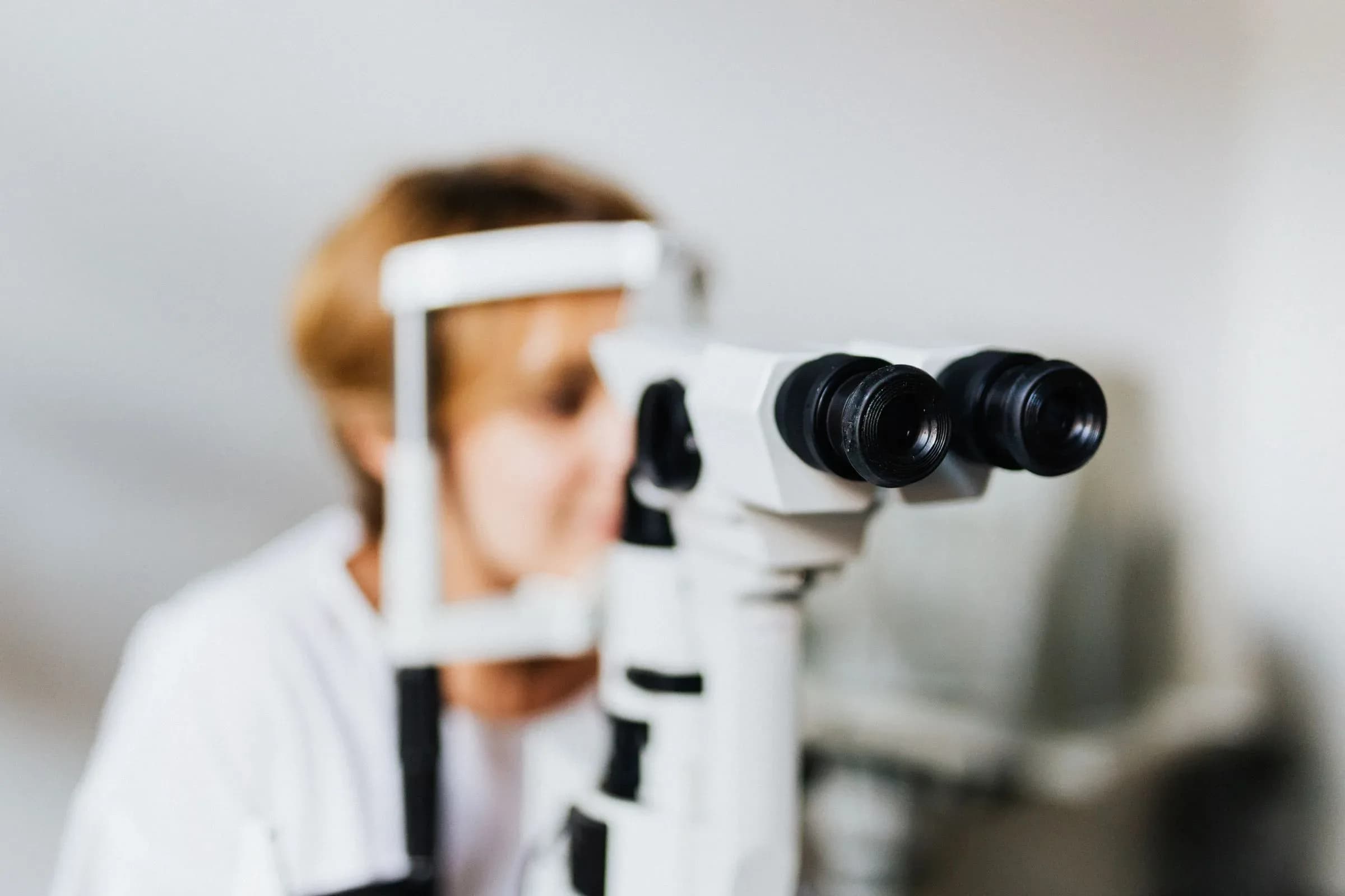 The eyepieces of a slit lamp in sharp focus, with a clinician blurred behind it.