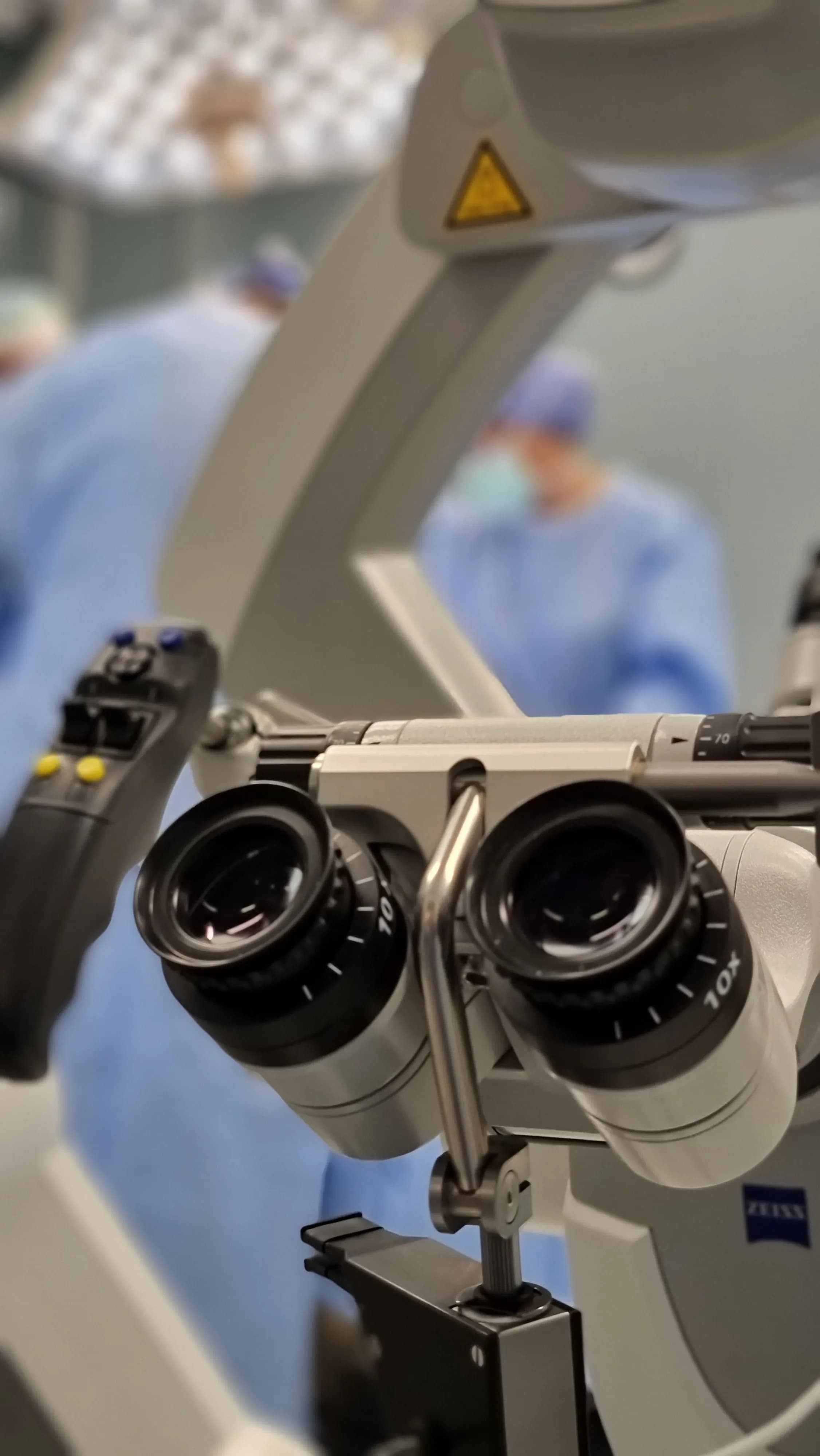The eyepieces of a surgical operating microscope in the foreground, with a gowned theatre team out of focus behind.