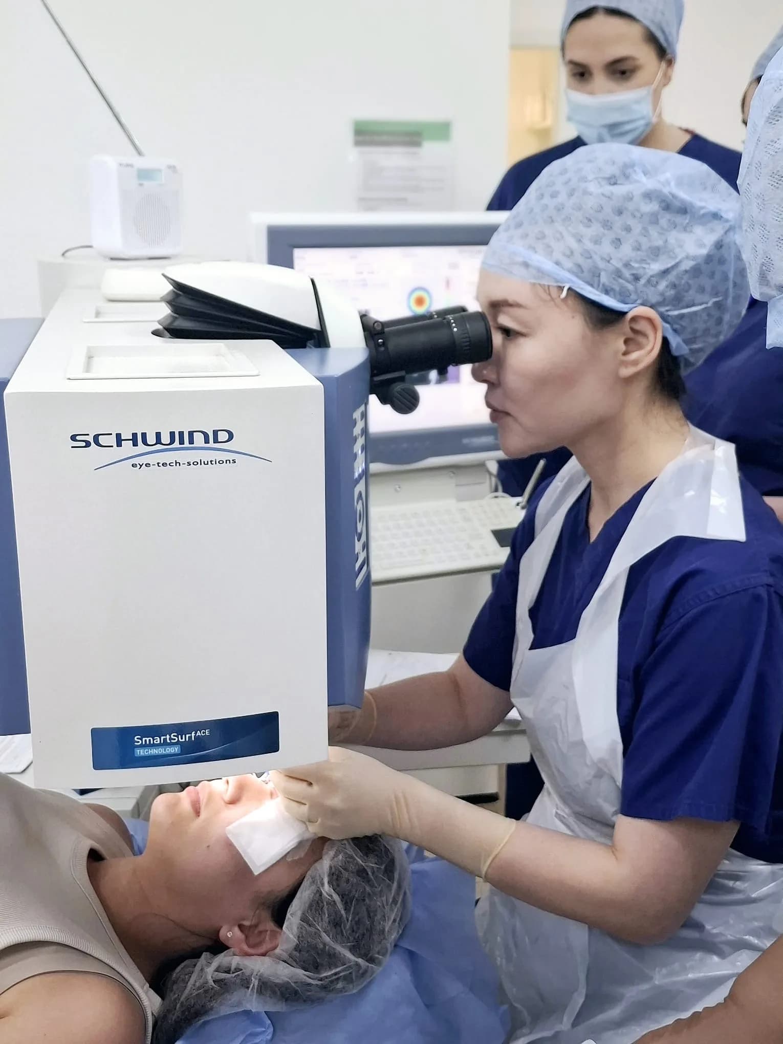 Ms Li Jiang in scrubs and a sterile apron looks through the microscope of an excimer laser while steadying a patient’s head; a colleague observes from behind.