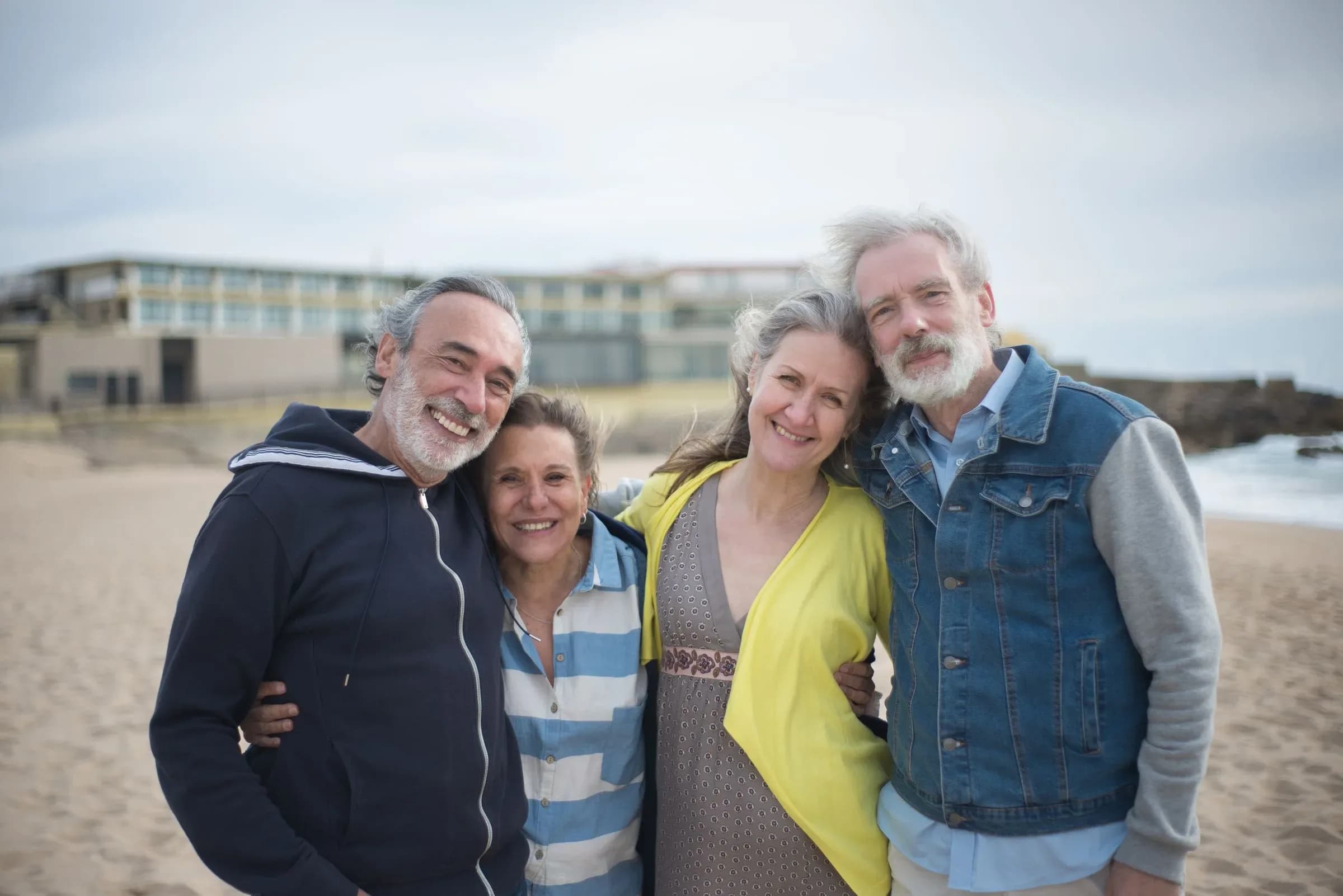 Four friends in their sixties standing together on a beach on an overcast day, smiling at the camera.