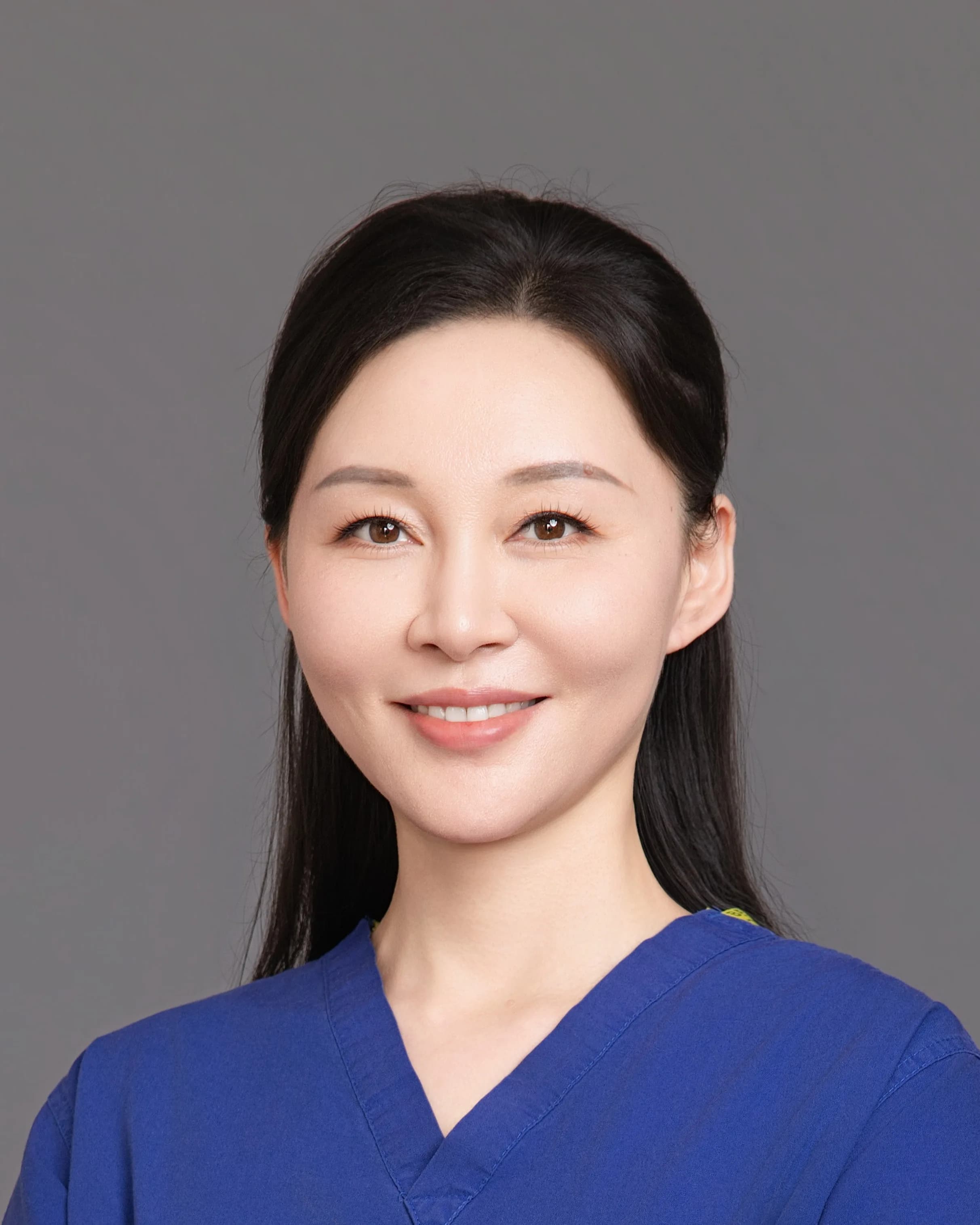 Ms Li Jiang, Consultant Ophthalmic Surgeon, in blue theatre scrubs with her arms folded, smiling, photographed against a plain grey background.