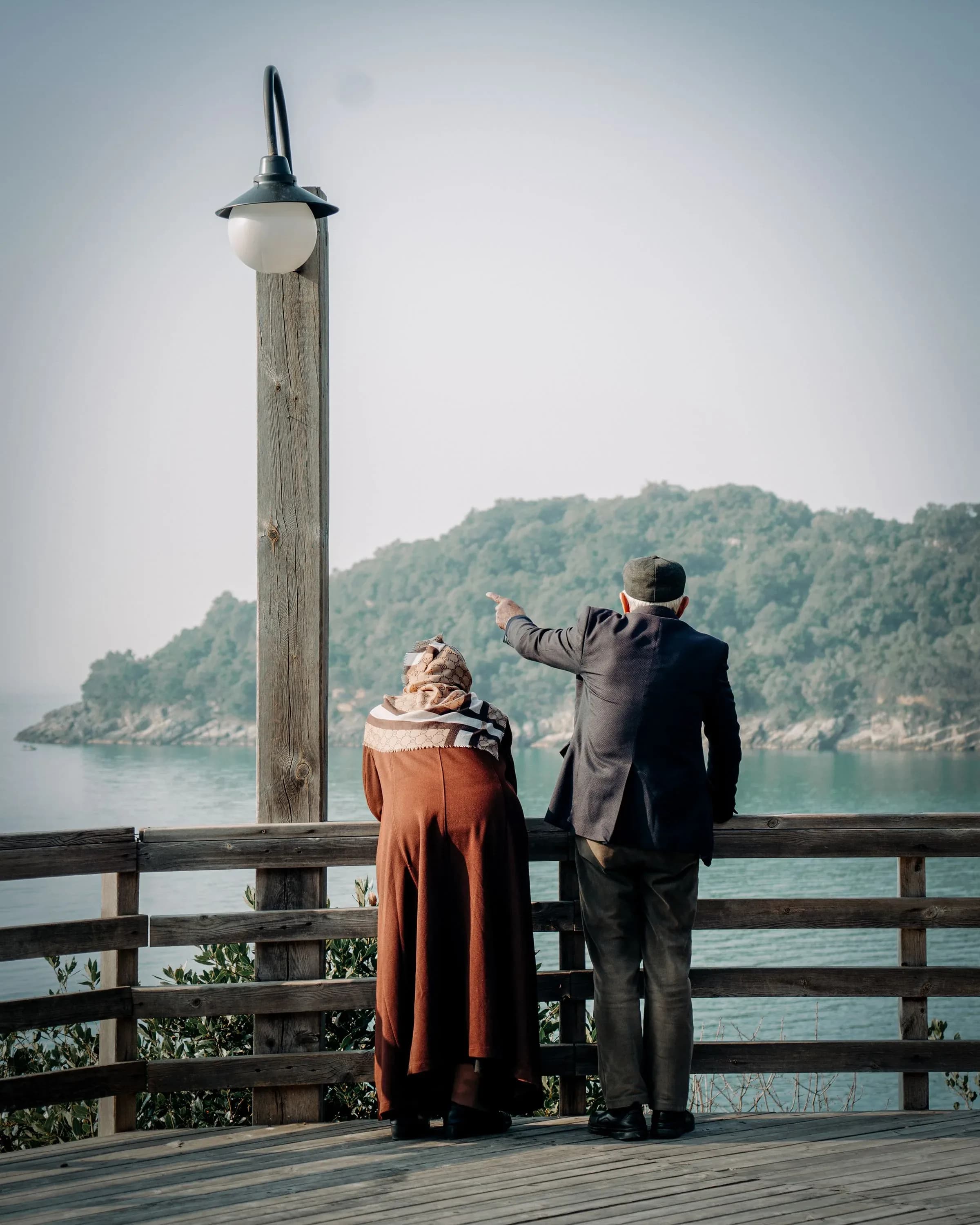 A couple standing at a wooden railing looking out across a bay, one pointing into the distance.