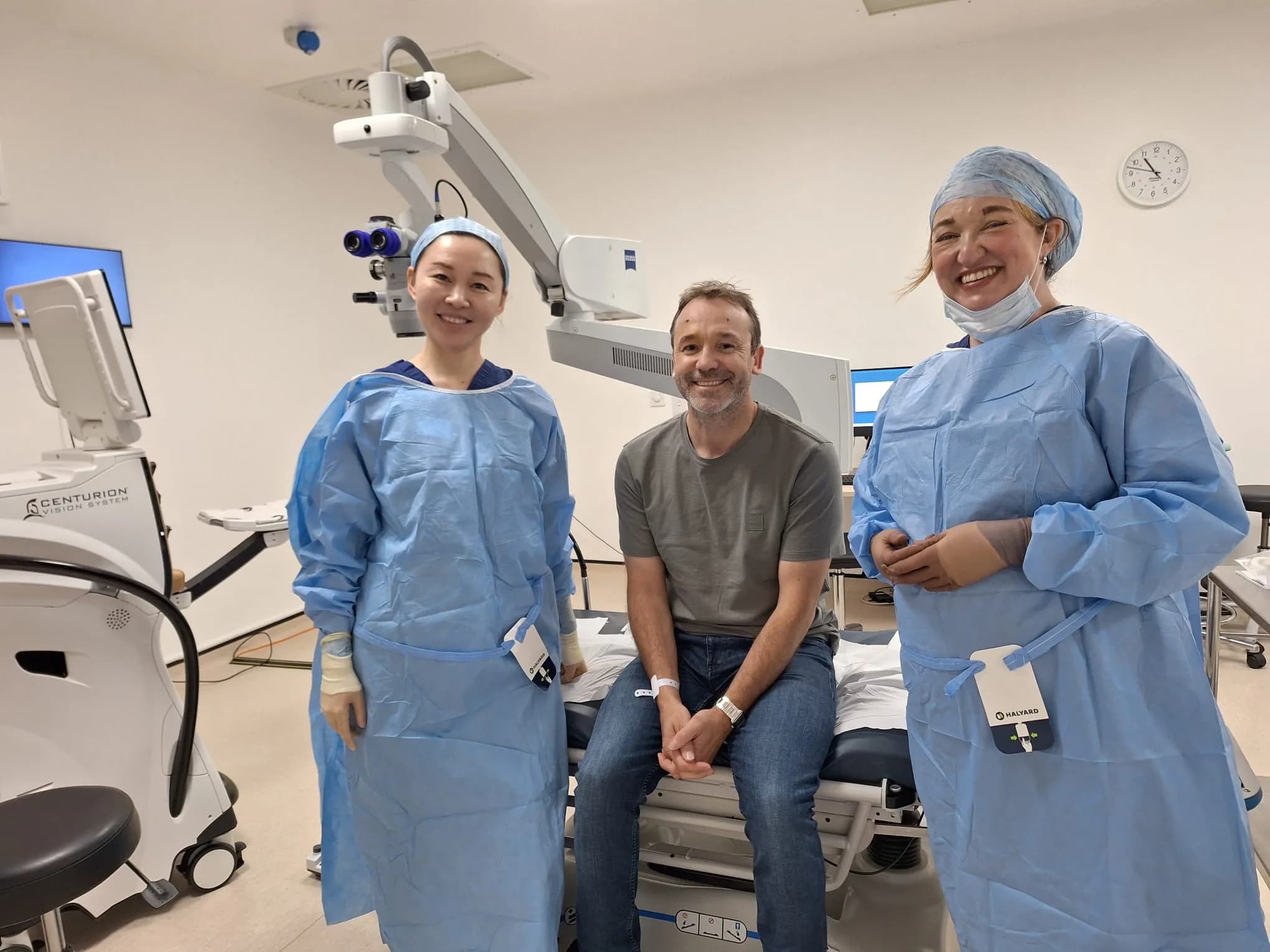 Ms Li Jiang and a colleague in sterile gowns and surgical caps stand either side of a patient seated on the treatment couch, with an operating microscope above them.