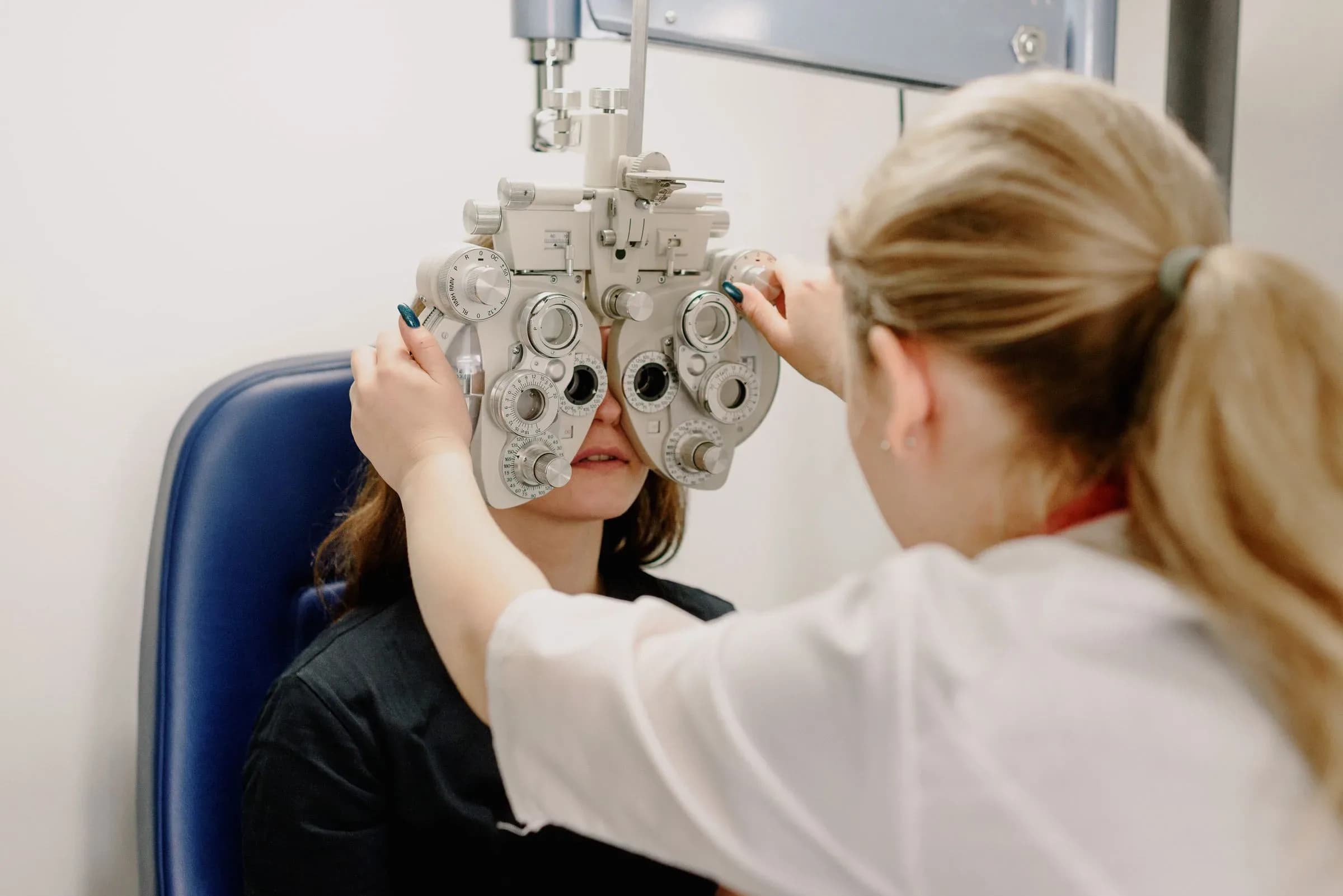 A clinician in a white coat adjusting a phoropter in front of a seated patient.