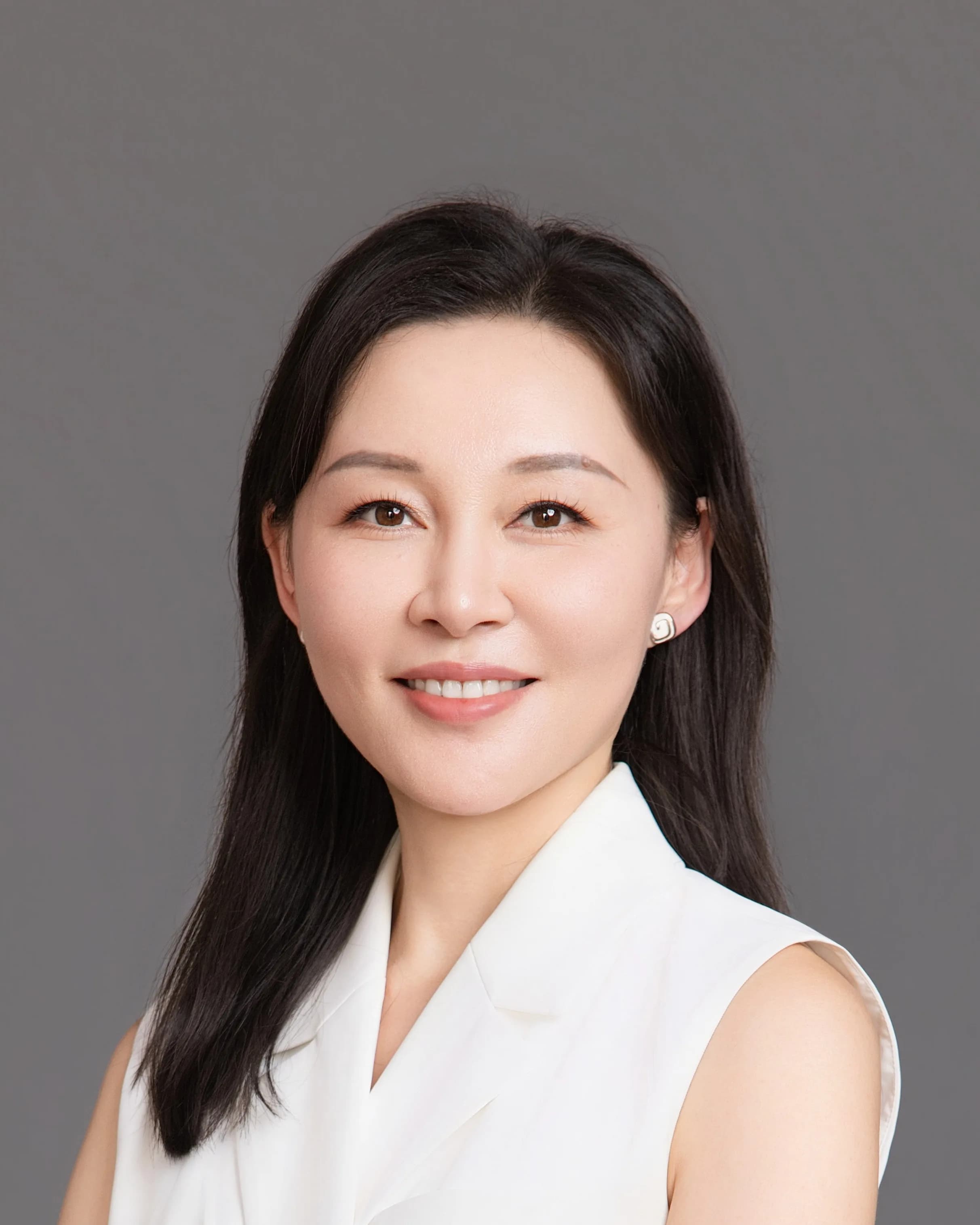 Ms Li Jiang, Consultant Ophthalmic Surgeon, in a white sleeveless jacket with her arms folded, smiling, photographed against a plain grey background.
