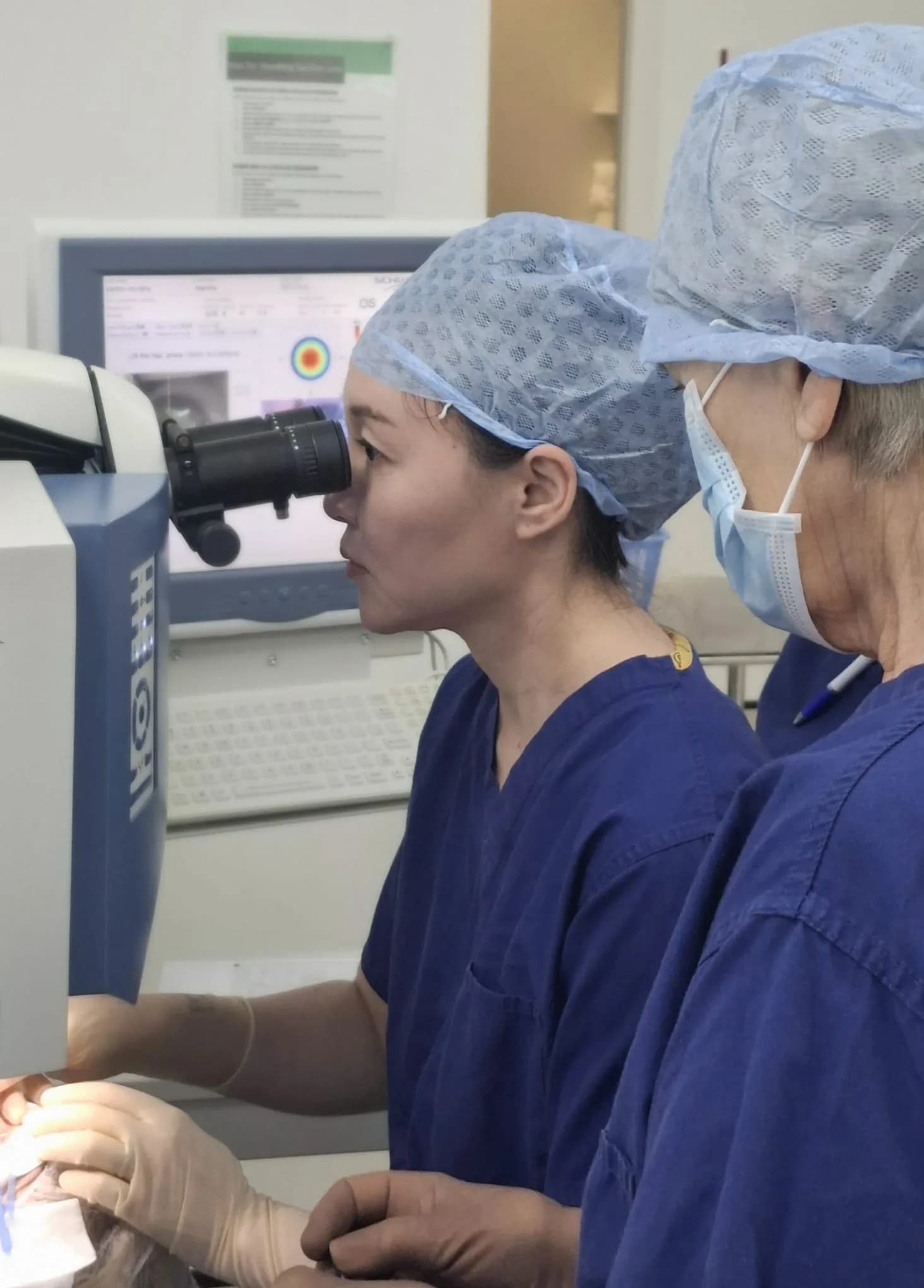 Ms Li Jiang in scrubs and a surgical cap looks into the eyepiece of a laser microscope while a masked colleague stands alongside; a screen behind them displays a corneal map.