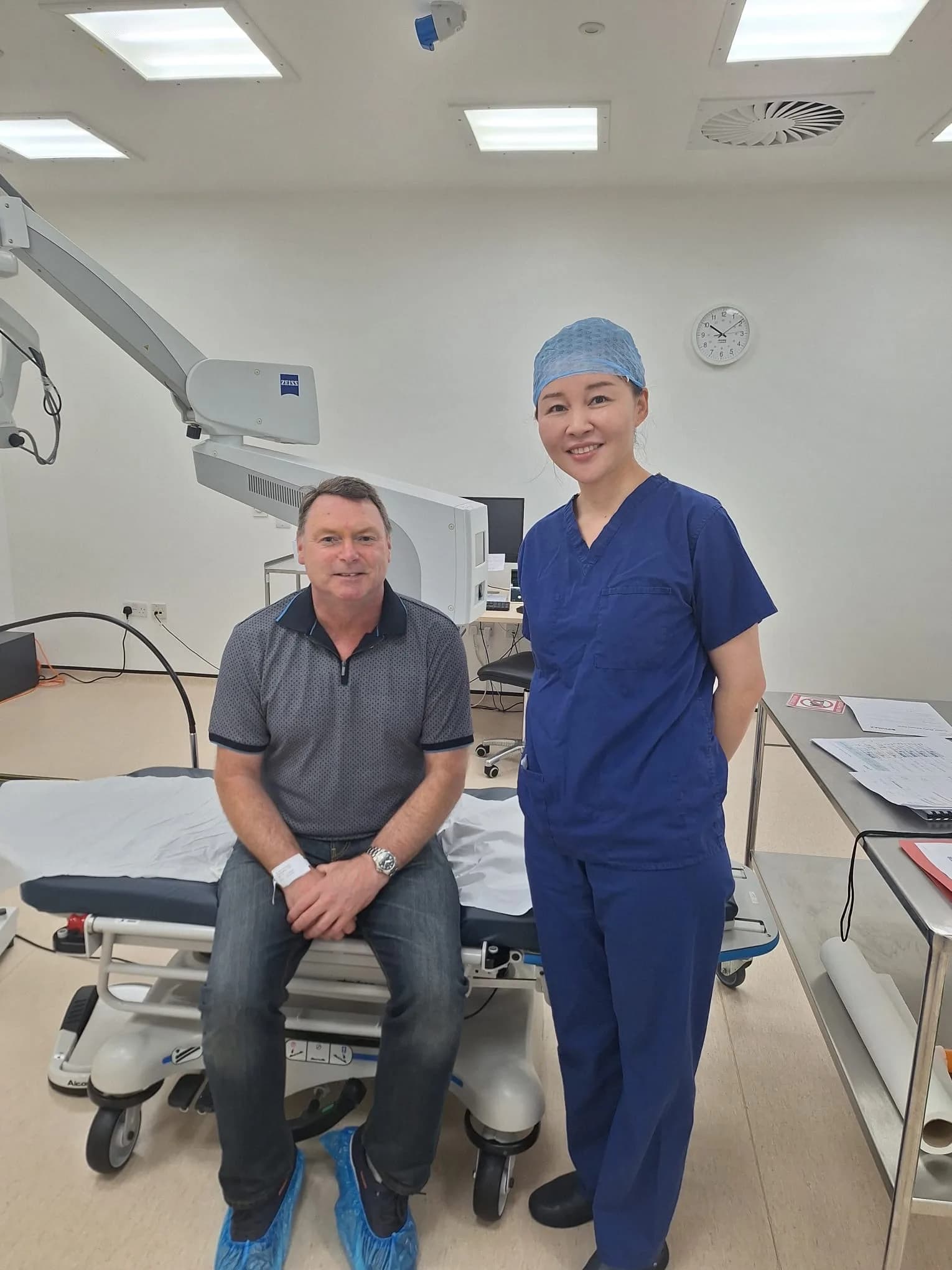 A patient in day clothes and shoe covers sits on the treatment couch beside Ms Li Jiang in scrubs, both smiling, with a laser system behind them.